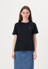 T-shirt in cotone nero con scollo rotondo e maniche corte, caratterizzato da un piccolo logo nella zona sinistra del petto. Abbinato a una gonna di jeans.