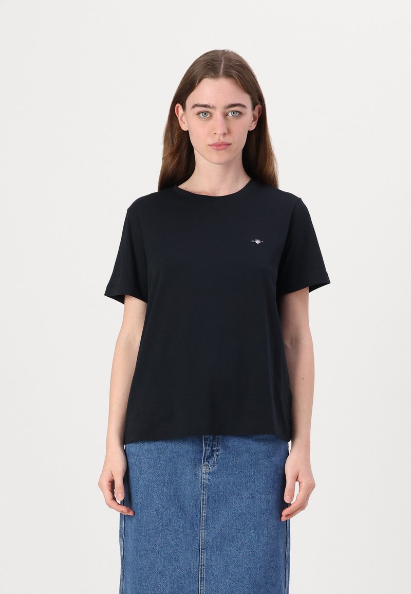 T-shirt in cotone nero con scollo rotondo e maniche corte, caratterizzato da un piccolo logo nella zona sinistra del petto. Abbinato a una gonna di jeans.
