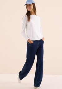 White long-sleeve top, dark blue wide-leg jeans with light stitching, white sneakers, and a light blue cap. Neutral background.