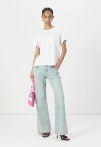 Tricou alb din bumbac cu mâneci scurte, purtat cu blugi din denim deschis, evazați. O mică geantă roz este ținută cu o mână, iar pe picioare sunt sandale negre.