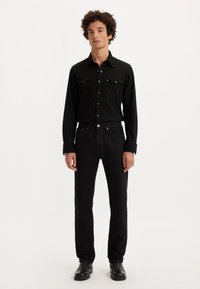 Mlad moški stoji, oblečen v črno srajco na gumb, črne kavbojke in črne škornje pred enotno svetlo ozadje.