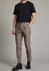 Svart kortärmad t-shirt, beige texturerade byxor med knappknäppning, och svarta snörskor, står mot en neutral grå bakgrund.