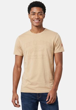 Jeune homme souriant portant un t-shirt graphique beige et un jean bleu, debout devant un fond uni clair.