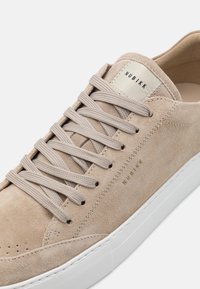 Beige velúrbőr sportcipő lapos bézs fűzőkkel, fehér gumitalppal és kis arany logóval az oldalon, letisztult, minimalista dizájnnal.