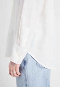 Chemise en lin blanche avec des manches longues légèrement froncées. Elle présente un poignet à bouton et un ourlet courbé, associée à un jean en denim bleu clair.