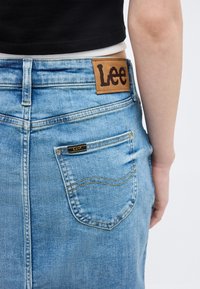 Κοντινό πλάνο πίσω μέρους φούστας από ανοιχτό μπλε τζιν με δερμάτινη ετικέτα της μάρκας Lee και τσέπη, φορεμένη από άτομο με μαύρο τοπ.