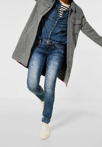 Cappotto grigio strutturato sopra una camicia di jeans, abbinato a jeans skinny azzurri e scarpe da ginnastica bianche. Caratterizzato da tasche e chiusure con bottoni.