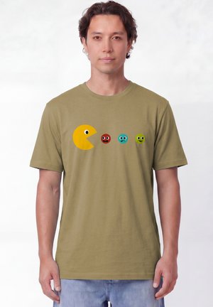 Homme portant un t-shirt vert olive avec un cercle jaune style Pac-Man poursuivant trois petits émoticônes colorés sur la poitrine.