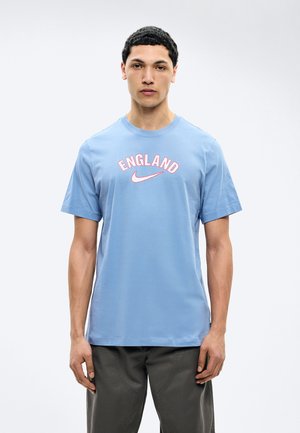 Jeune homme aux cheveux courts et bouclés portant un t-shirt Nike "England" bleu clair et un pantalon foncé, debout devant un fond blanc uni.
