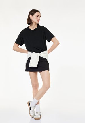 Young woman standing, wearing black t-shirt and skirt, white sweater tied around waist, white socks, and white sneakers with gum soles.