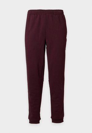 Uomo in piedi che indossa una t-shirt bianca a maniche corte, pantaloni da jogging color bordeaux e scarpe da ginnastica bianche su uno sfondo chiaro e uniforme.