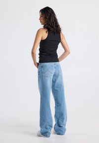 Woman with wavy dark hair wearing a black sleeveless top and light blue wide-leg jeans, standing with hands on hips facing away.