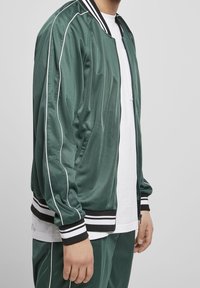 Grüne Satin-Bomberjacke mit weißen Paspeln, schwarz-weißen gestreiften Rippbündchen und zwei seitlichen Taschen. Über einem weißen T-Shirt getragen.