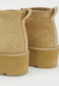 Klasiska tan krāsas zamšādas potītes kurpes ar gludu tekstūru, noapaļotu formu un vieglu gumijas zoli. Iezīmē redzamas šuves un neliela cilpa uz papēža.