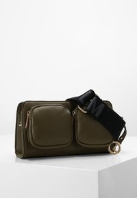 Olijfgroene leren tas met twee vooroplopende ritszakken, bevestigde zwarte band met logo, hardware in goudtint, gladde textuur.