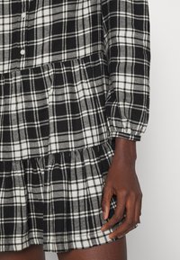 Svart och vit rutad klänning med långa ärmar, knäppning fram, samlad midja och flerfaldig volang nederkant. Mjukt tyg, avslappnad design.