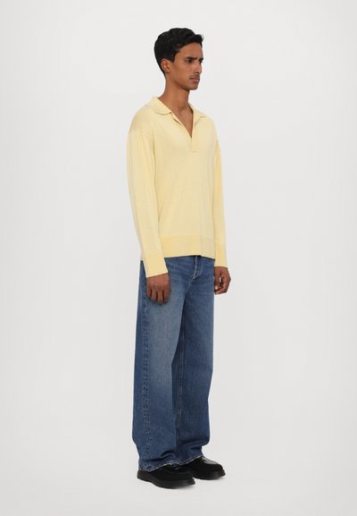 Gelbes Langarm-Polohemd mit V-Ausschnitt, gerippten Bündchen und lockerer Passform, kombiniert mit lockeren blauen Jeans aus Denim und schwarzen Schuhen.