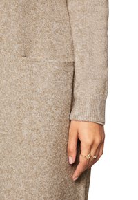 Béžový pletený svetr s detailním pohledem na ruku u kapsy. Tkanina má strukturovaný povrch a rukáv obsahuje žebrované detaily.