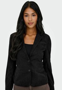 Blazer en velours côtelé noir à deux boutons, avec un col cranté, deux poches avant et un tissu texturé. Porté par-dessus un haut noir.