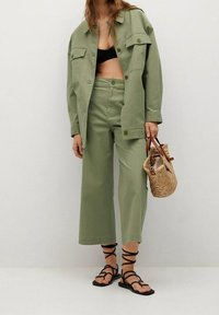 Zielona khaki kurtka z dwiema kieszeniami na piersi, dopasowane szerokie spodnie w stylu cropped, czarne sandały z paskami oraz pleciona brązowa torebka.