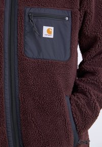 Brown fleece jacket with a black nylon chest pocket featuring a zipper and logo. The fabric has a textured, fluffy appearance.