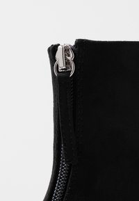 Bottine en daim noir avec une fermeture éclair argentée sur le côté, une surface texturée et un design épuré et arrondi pour une apparence élégante.