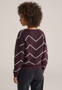 Burgunderfarbener Pullover mit weißem Zickzackmuster, aus weichem Material gefertigt. Verfügt über einen weiten Ausschnitt und gerippte Bündchen, kombiniert mit dunklen Jeans.