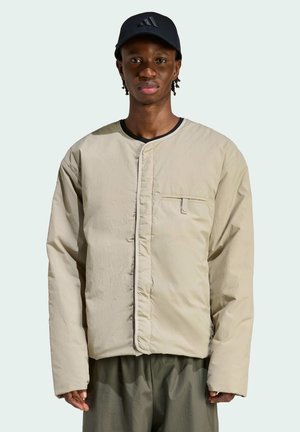 Młody mężczyzna w beżowej, bezkołnierzowej, ocieplanej kurtce, oliwkowych spodniach i czarnej czapce Adidas, stojący na tle jasnego, jednolitego tła.