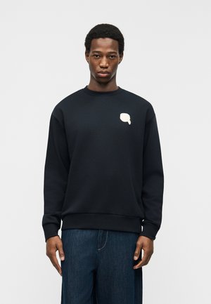 Young man wearing a black sweatshirt with a small white speech bubble design on the left chest, paired with dark blue jeans.