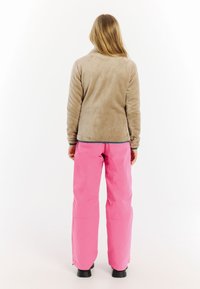 Bruine fleece jas en lichtroze waterdichte broek. De jas heeft een zachte textuur; de broek is gestroomlijnd met ritsen bij de enkels.