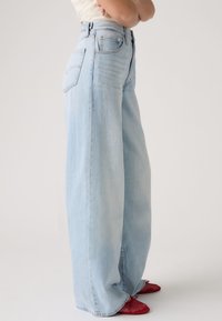 Personne portant un jean large bleu clair et des chaussures plates rouges texturées, debout les bras croisés devant un fond clair uni.