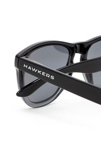 Musta aurinkolasit kiiltävällä viimeistelyllä, joissa tummat linssit ja valkoisin kirjaimin "HAWKERS" painetut sankat. Pyöreä kehys.