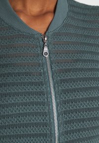 Getextureerde teal ritssluitingjas met een geribbelde kraag, voorzien van een zilveren rits en een horizontaal streeppatroon over de hele stof.