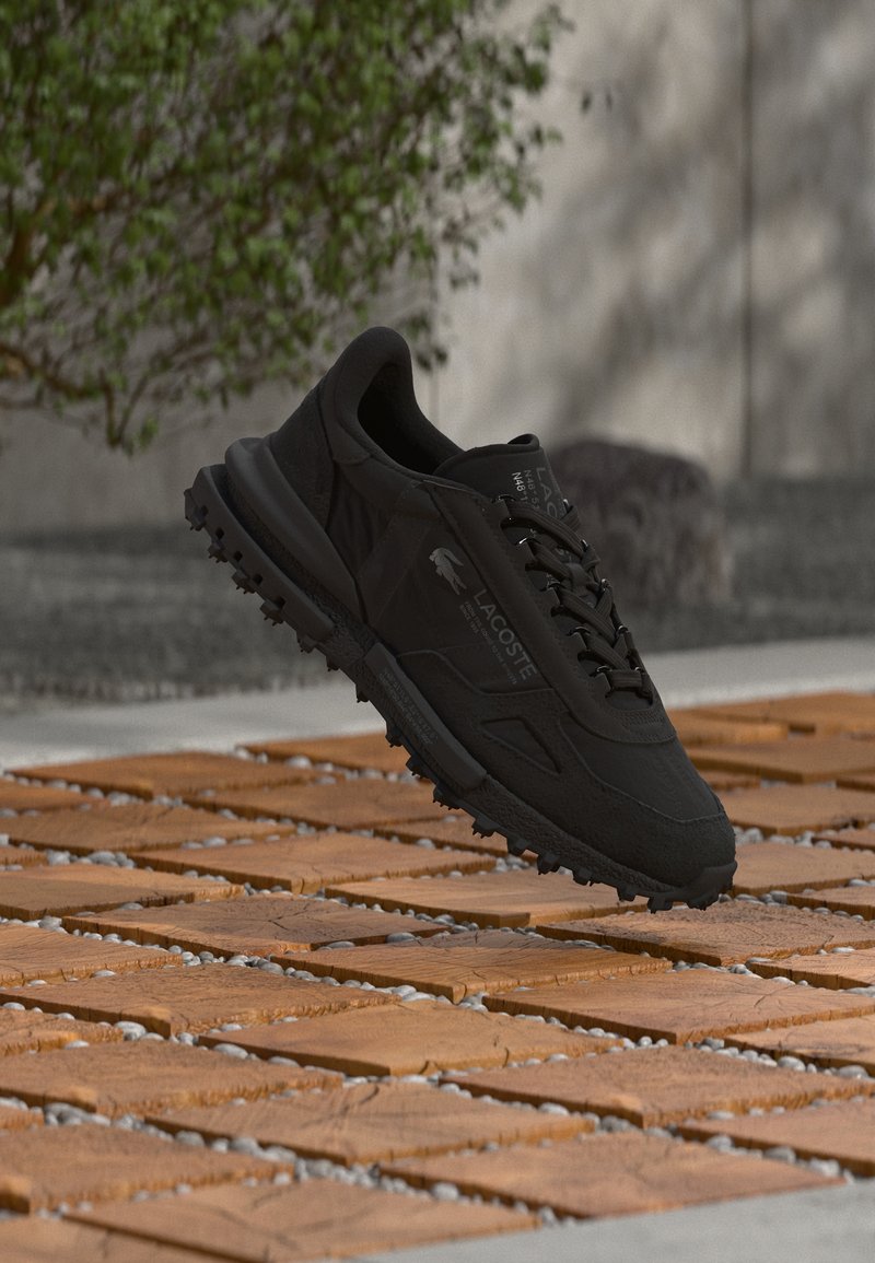 Baskets noires Lacoste avec semelle texturée flottant au-dessus d'une surface en carreaux de bois en plein air, près d'un arbre flou et d'un mur en arrière-plan.