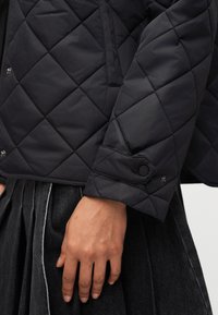 Κοντινό πλάνο ενός χεριού που ξεκουράζεται στο πλάι, φοράει μαύρο καπιτονέ μπουφάν με κουμπιά τύπου snap και σκουρόχρωμη πλισαριστή φούστα.