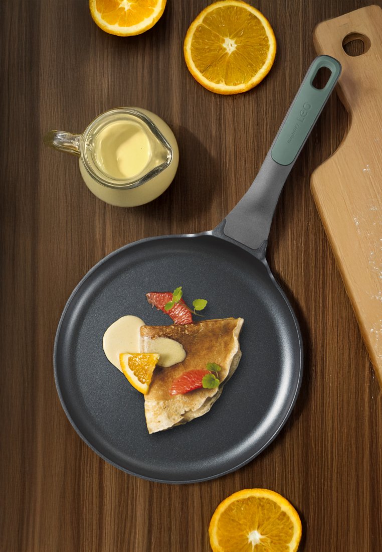 Eine beschichtete Pfanne mit einem grauen Körper und einem grünen Griff, die einen Pfannkuchen mit einer Garnierung aus Orange, Grapefruit, Minze und Sahne auf einer Holzoberfläche hält.