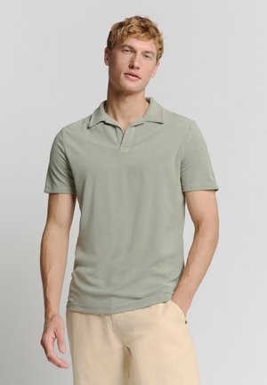 Kurzarm-Poloshirt aus hellgrünem Stoff mit weicher Textur, offener Kragen und geradem Saum, kombiniert mit hellbeigen Hosen.