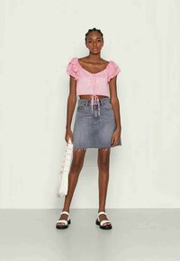 Top curto em rosa claro com mangas com babados, combinado com uma saia lápis de denim azul. A modelo segura uma bolsa branca e usa sandálias pretas.