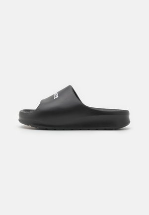 Sandalias negras sin hebilla con superficie de goma suave, punta redondeada, suela texturizada y logo blanco en la correa.