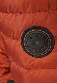 Detalje af en orange pufferjakke med et rundt sort tekstil-patch, der har prægede bogstaver "CMS SPORT" og et logo, som fremhæver en struktureret overflade.