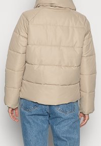 Beżowa kurtka puchowa z kapturem, z pikowanym wzorem i ściągaczami, w połączeniu z jasnoniebieskimi dżinsami.