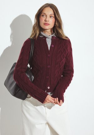 Burgundy cable-knit cardigan with a fitted design, round neckline, purple buttons, and ribbed cuffs; paired with a gray top and white pants.