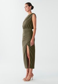 Olijfgroene, geribbelde midi-jurk met een ontwerp met één schouder en een split aan de zijkant. Heeft een getailleerde taille en een rechte silhouet. Draagt bijpassende sandalen met bandjes.