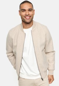 Beige bomber jacket with ribbed collar and cuffs, zip closure, and soft texture, worn over a white t-shirt.