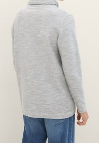Grauer Langarmpullover mit strukturiertem Rippmuster, gefertigt mit einem hohen Kragen und in lockerer Passform, kombiniert mit blauen Jeans.
