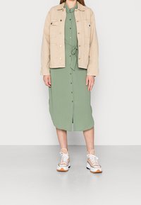 Beige denimjacka över en grön skjortklänning med midjeband. Vita sneakers med orangea detaljer. Enkel och funktionell design.