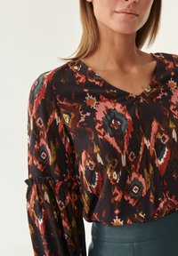 Blouse à motifs avec un col en V, fond sombre orné de motifs rouges, pêche et bleu sarcelle, et manches froncées. Texture du tissu lisse.
