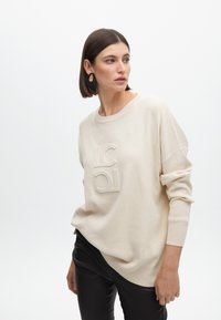 Felpa lavorata a maglia beige con logo in rilievo. Presenta un collo a giro e una vestibilità comoda, abbinata a pantaloni in pelle nera. Texture liscia.