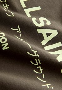 Tela de algodón en marrón oscuro con texto en amarillo claro y elementos gráficos en negrita. Textura suave, ligeramente arrugada, que muestra detalles del diseño.
