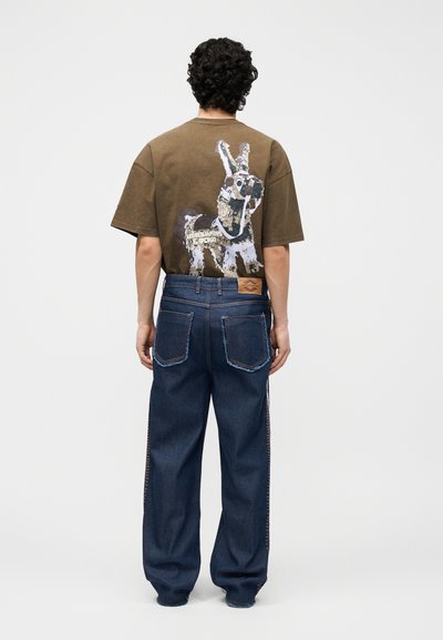 Άνδρας που φαίνεται από πίσω, φοράει καφέ κοντομάνικο μπλουζάκι με γραφικό ενός γαϊδάρου και σκούρο μπλε παντελόνι με φαρδιά πόδια και μεγάλες πίσω τσέπες.